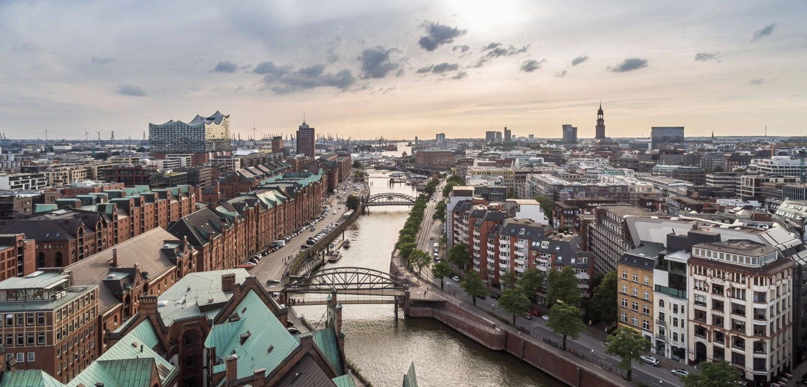 wir blicken von oben auf die Kanäle der Hamburger ALtstadt, auf roten Backstein und im HIntergrund das gebäude der Elbphilharmonie mit seinem wellenförmigen Dach. Rehct im BIld der Turm der Michaeliskirche.
